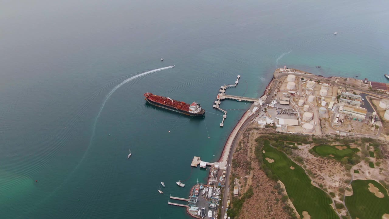 Aerial frame in La Paz showing the red cargo ship left of the industrial dock, pale turquoise shallows along the shoreline, small boats near the marina, golf course terrain, and no visible people.