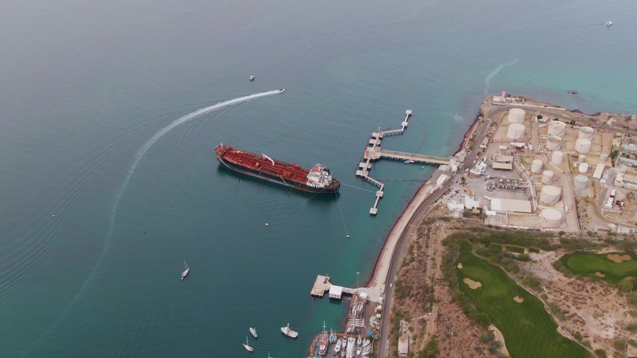 High drone-like view of La Paz harbor with the red cargo ship centered left, a long industrial pier to the right, storage tanks onshore, a fading speedboat wake near the top water, and no visible people.