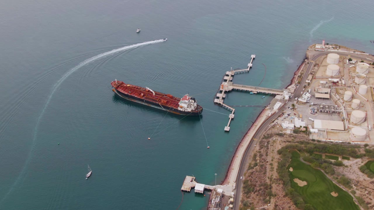 Closer aerial view of a red cargo ship near shore facilities and a curved boat wake.