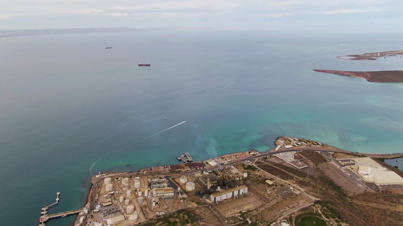 Opening aerial frame over La Paz, Baja California Sur showing tanks, docks, low industrial buildings, turquoise shallows, distant ships, and no visible people.