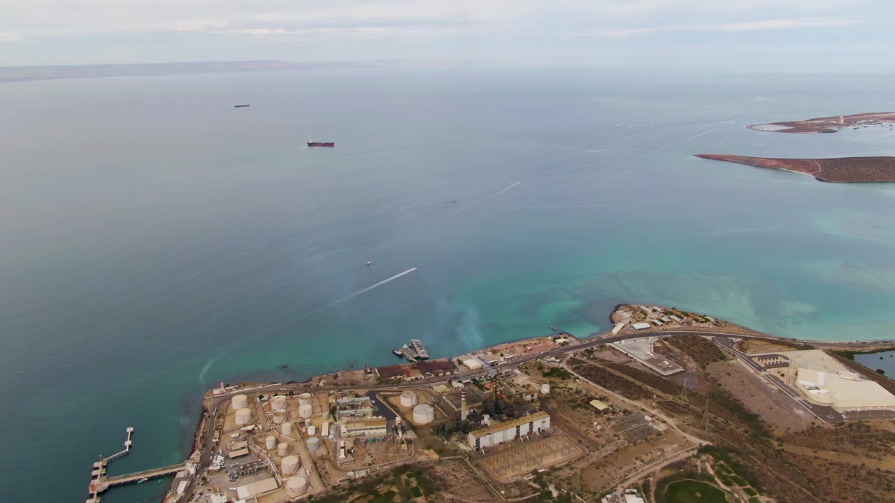 Aerial view in La Paz, Baja California Sur with the industrial site lower in frame, turquoise water offshore, two distant ships, and no visible people.