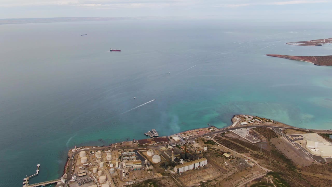 Mid-clip aerial over La Paz, Baja California Sur showing the shoreline facility, pale coastal shallows, a ship offshore, a faint wake, and no visible people.
