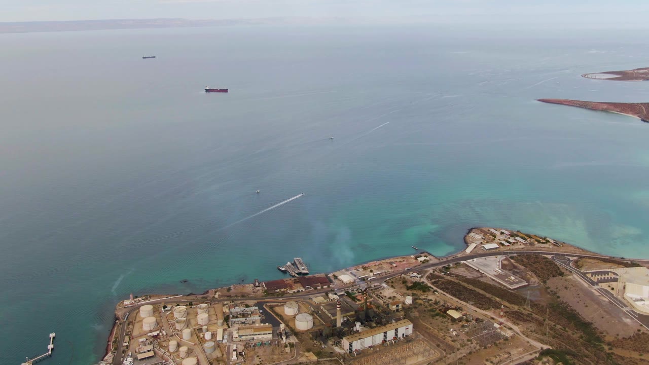 Later aerial frame in La Paz, Baja California Sur with more open water in view, a small white wake crossing the sea, shoreline tanks below, and no visible people.
