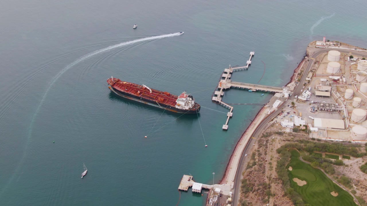 Aerial frame over La Paz harbor with the red cargo ship held near center-left, a curved white wake sweeping across the upper water, industrial tanks at right, and no visible people.