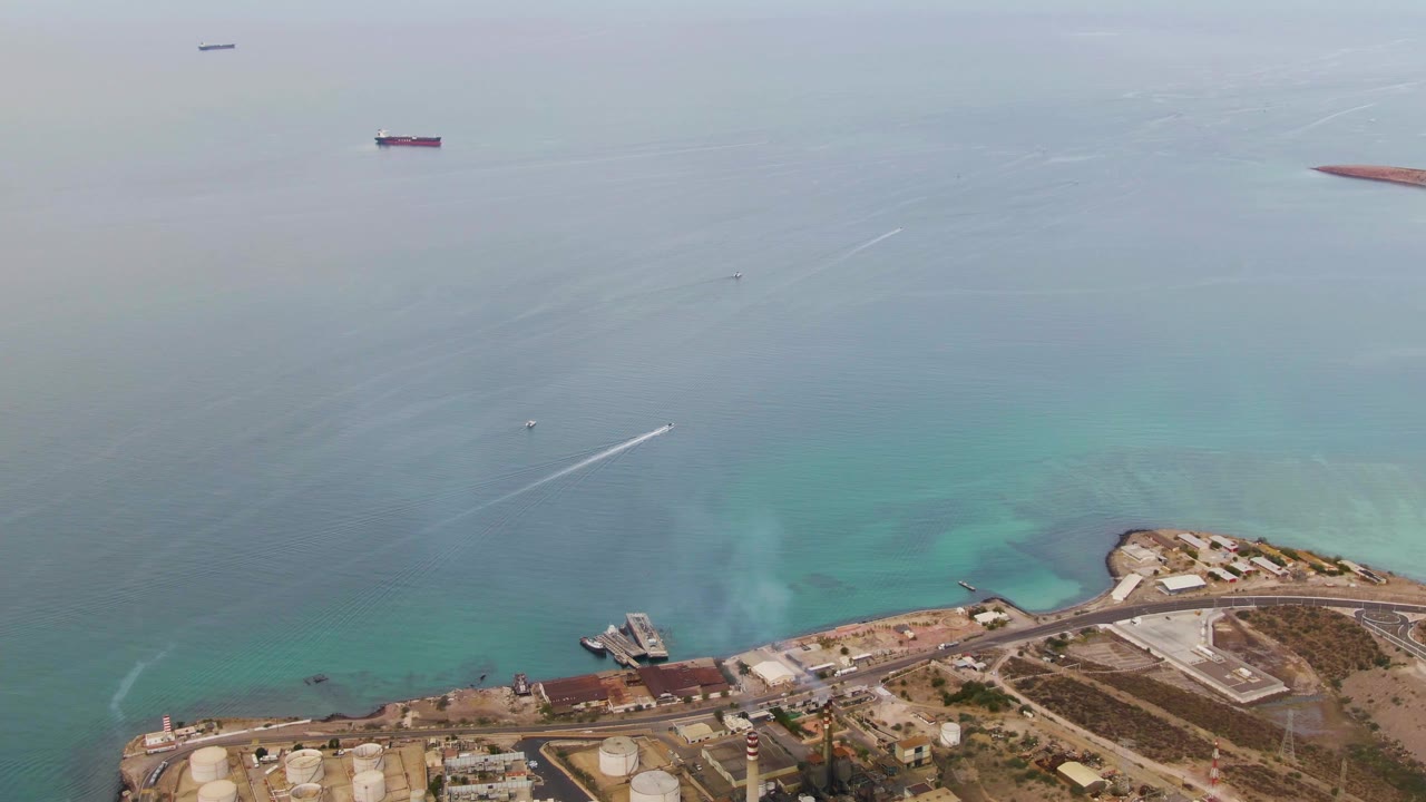 Aerial view over La Paz, Baja California Sur, with industrial shoreline at the bottom, turquoise coastal water, offshore ships, a small boat wake, and no visible people.