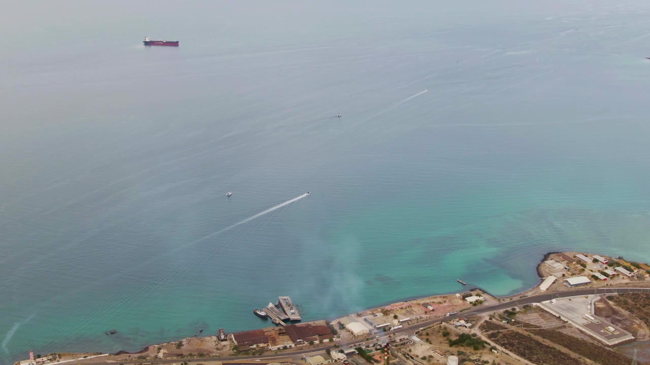 High aerial frame from La Paz, Baja California Sur, with turquoise shallows, faint smoke or haze near the industrial shoreline, offshore vessels, a bright boat wake, and no visible people.