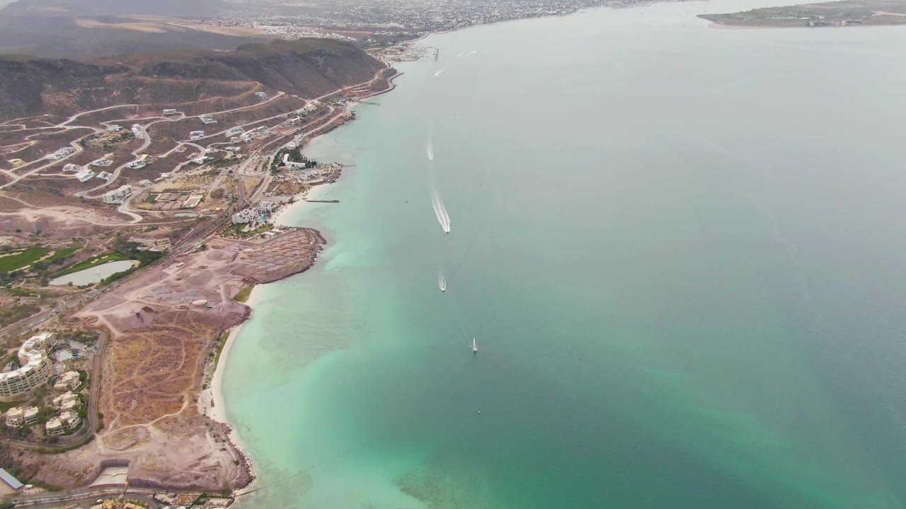 Aerial frame from La Paz with pale turquoise shallows along the beach, dry tan land and hillside development to the left, boats leaving white wakes offshore, and no visible people.