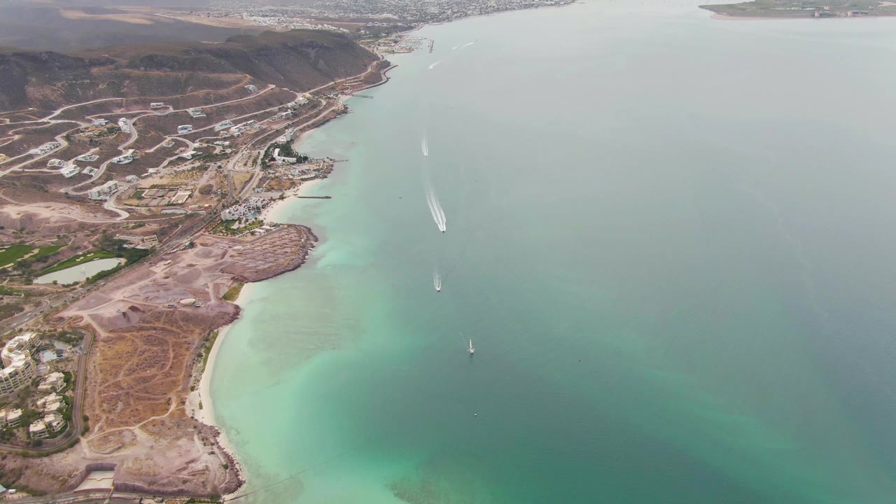 Aerial view in La Paz showing a sandy beach below dry coastal terrain, hillside roads and structures in the background, several boats spread across blue-green water, and no visible people.