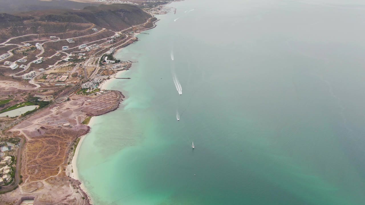 Aerial frame over La Paz with the lower beach and dry foreground more visible, turquoise shallows hugging the shore, hillside development beyond, boats offshore, and no visible people.