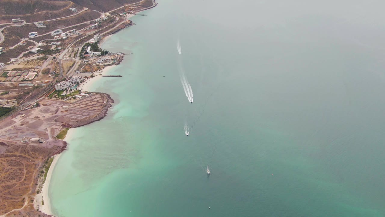 Aerial view over La Paz, Baja California Sur coastline showing turquoise shallows, dry shoreline, small boats, and a speedboat wake near the upper right, with no visible people.