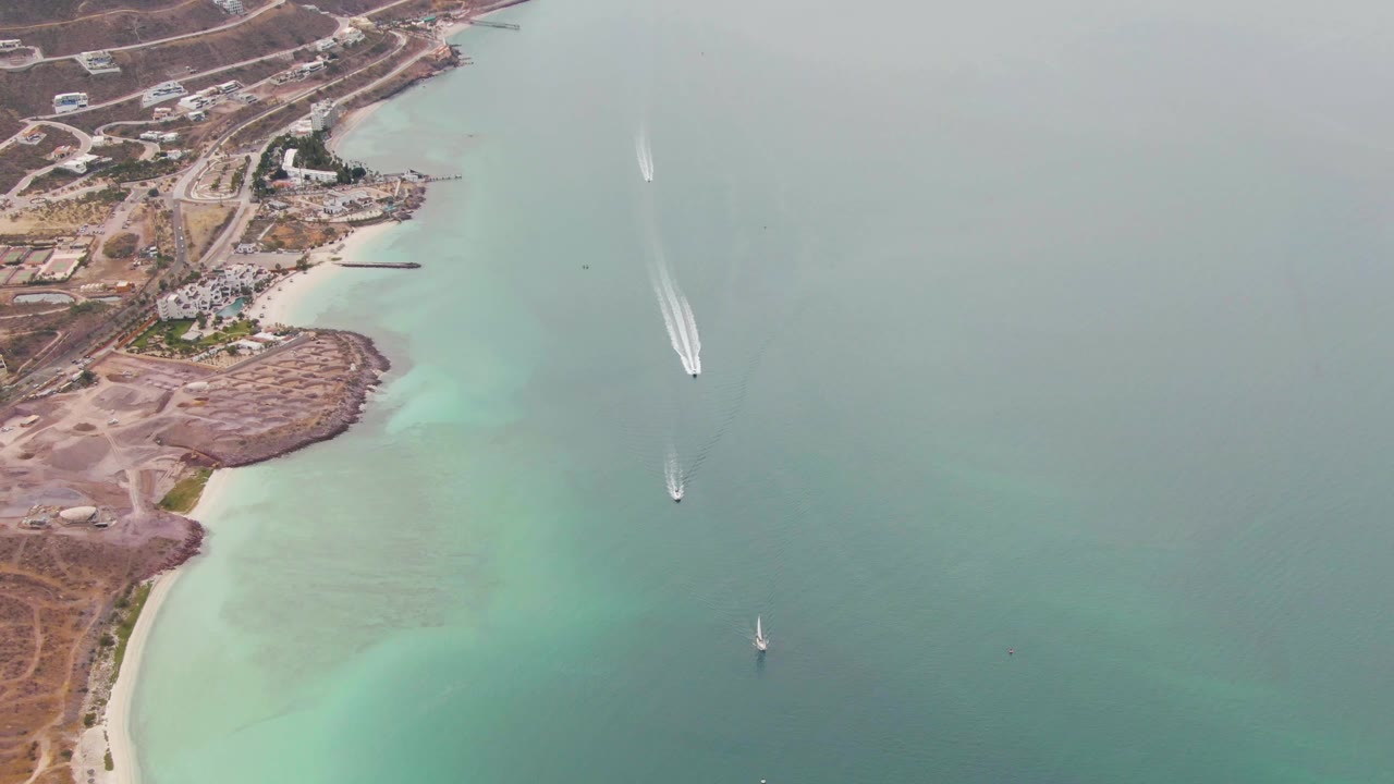 Aerial frame from La Paz, Baja California Sur with a white speedboat wake farther out, a small sailboat below it, pale turquoise water near shore, and no visible people.
