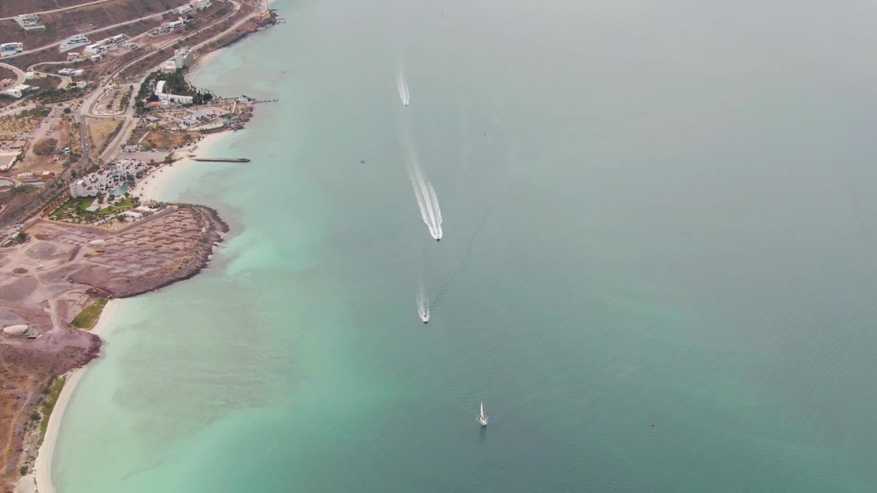 La Paz, Baja California Sur aerial still showing shoreline development at upper left, a long white boat wake in open water, a sailboat nearer the center-right, and no visible people.