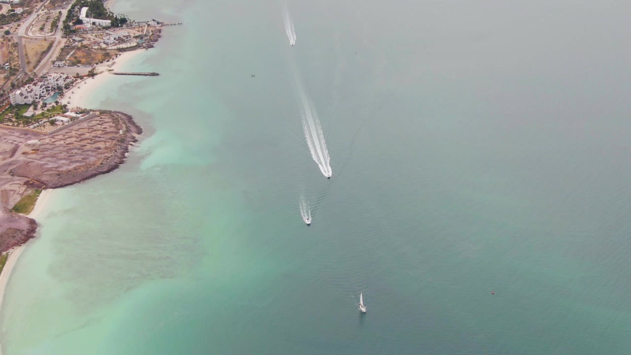 Aerial frame over La Paz, Baja California Sur, showing boats crossing blue-green coastal water near a rocky point, sandy beach, roads, buildings, and no visible people.