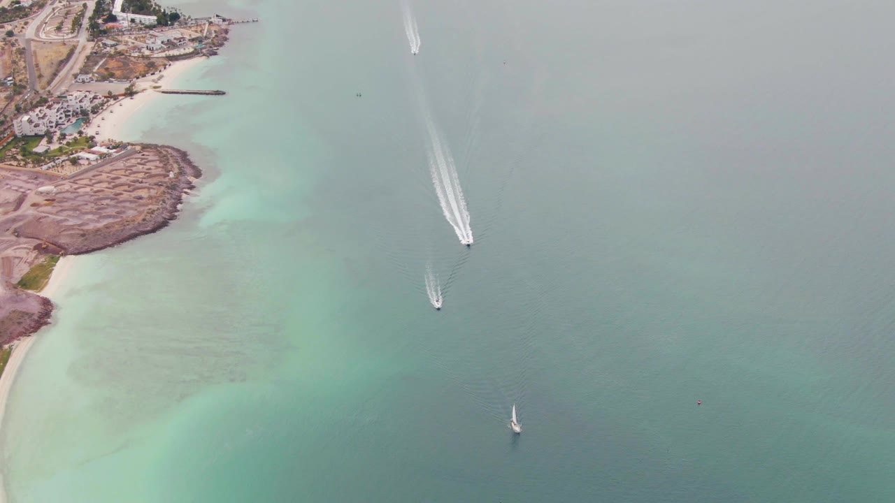 Aerial frame over La Paz coastal water with two moving boats, long white wake trails, pale shallows along the left shoreline, and no visible people.