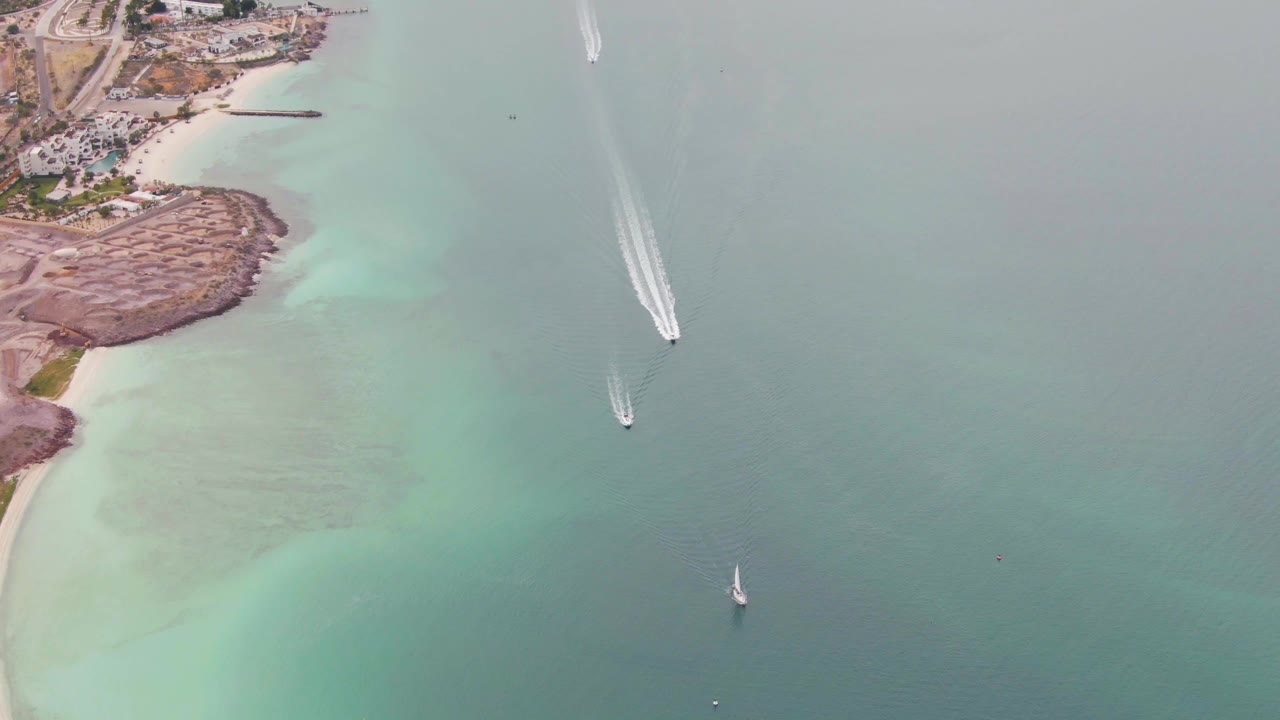 Aerial frame in La Paz, Baja California Sur, with a rocky shoreline at left, pale turquoise shallows, boats separated across the water, wake trails, and no visible people.