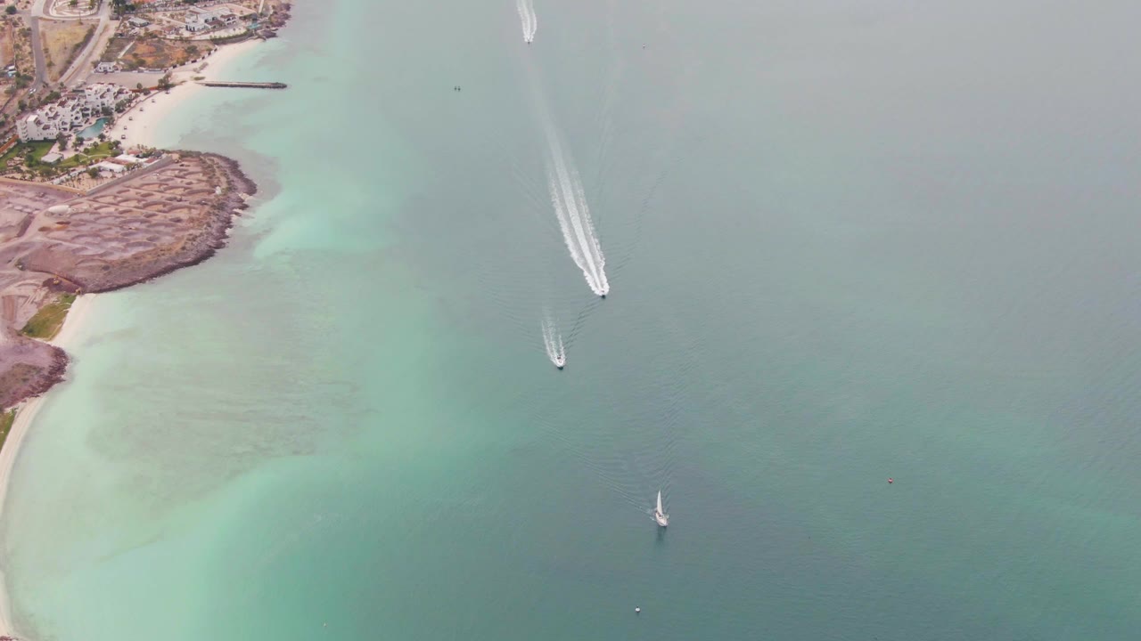Drone view of turquoise shallows, sandy beach, rocky coast, and boats with white wakes.