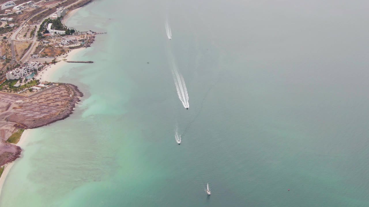 Aerial frame over La Paz, Baja California Sur, with boats cutting across green-blue coastal water beside a sandy beach, rocky shore, scattered buildings, and no visible people.
