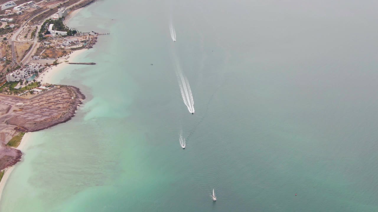 Boats move through calm coastal water near a beach and rocky shoreline.