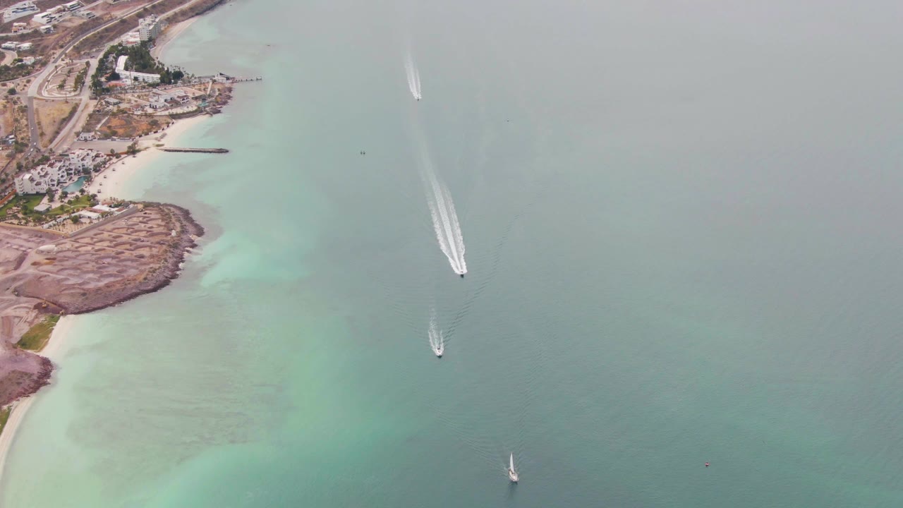 Drone-like overhead frame in La Paz with boat wakes shifting farther across the open water, a small sailboat lower in frame, rocky shore, beach, buildings, and no visible people.