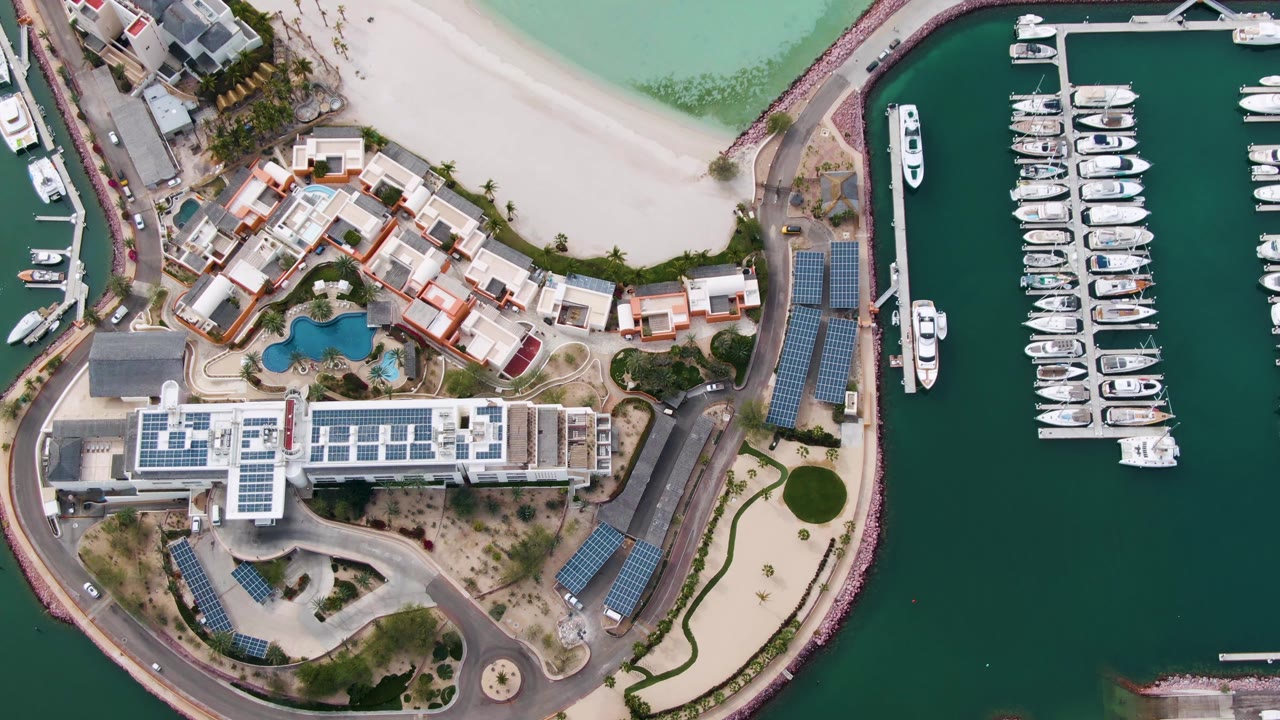 Opening overhead frame of a La Paz, Baja California Sur marina with docked white boats, a pale beach, waterfront buildings, roads, and no visible people.