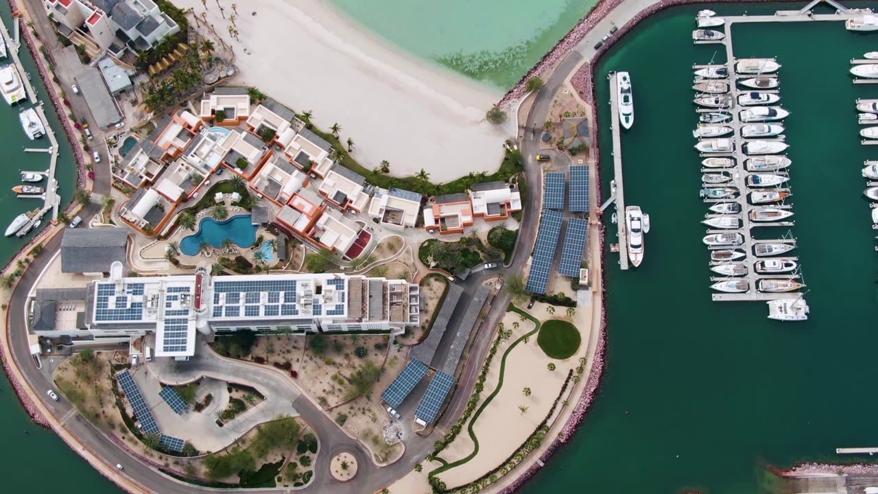 Second overhead moment in La Paz showing the beach curve, clustered waterfront buildings, docked marina boats, darker harbor water, and no visible people.
