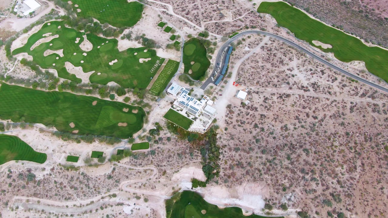 A later aerial frame shows more of the lower fairway edge and clubhouse area in La Paz, with dry scrubland filling the right side and no visible people.
