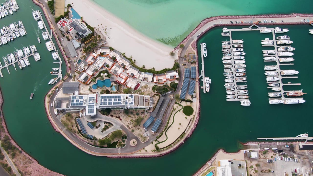 Top-down aerial frame of a La Paz marina with white yachts in slips, island buildings, curved roadway, pale beach, turquoise shallows, and no visible people.
