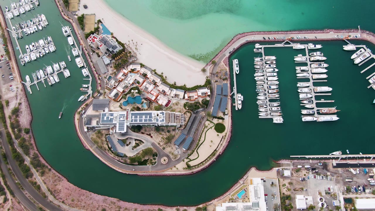 Overhead La Paz marina frame with rows of white boats in the enclosed basin, a curved rock shoreline around island buildings, road and parking below, and no visible people.