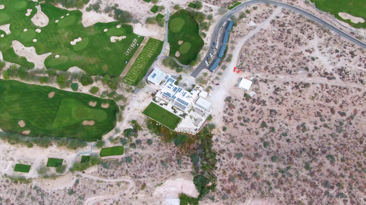Aerial frame in La Paz, Baja California Sur, looking down on fairways, small sand traps, a roadside edge, practice areas, dry scrubland, and no visible people.