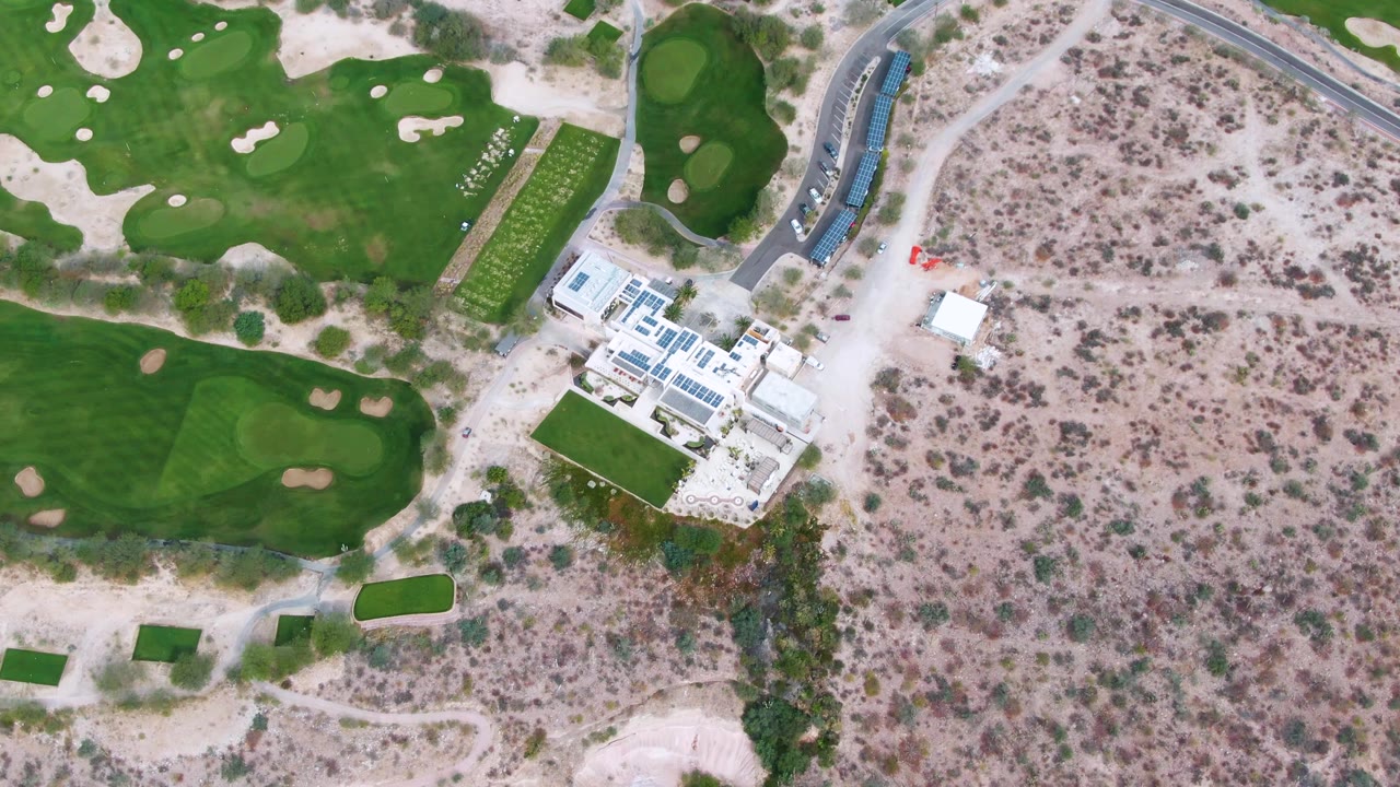 Near-vertical overhead view in La Paz, Baja California Sur, with a cluster of white buildings between green golf turf and scrubby desert ground, with no visible people.