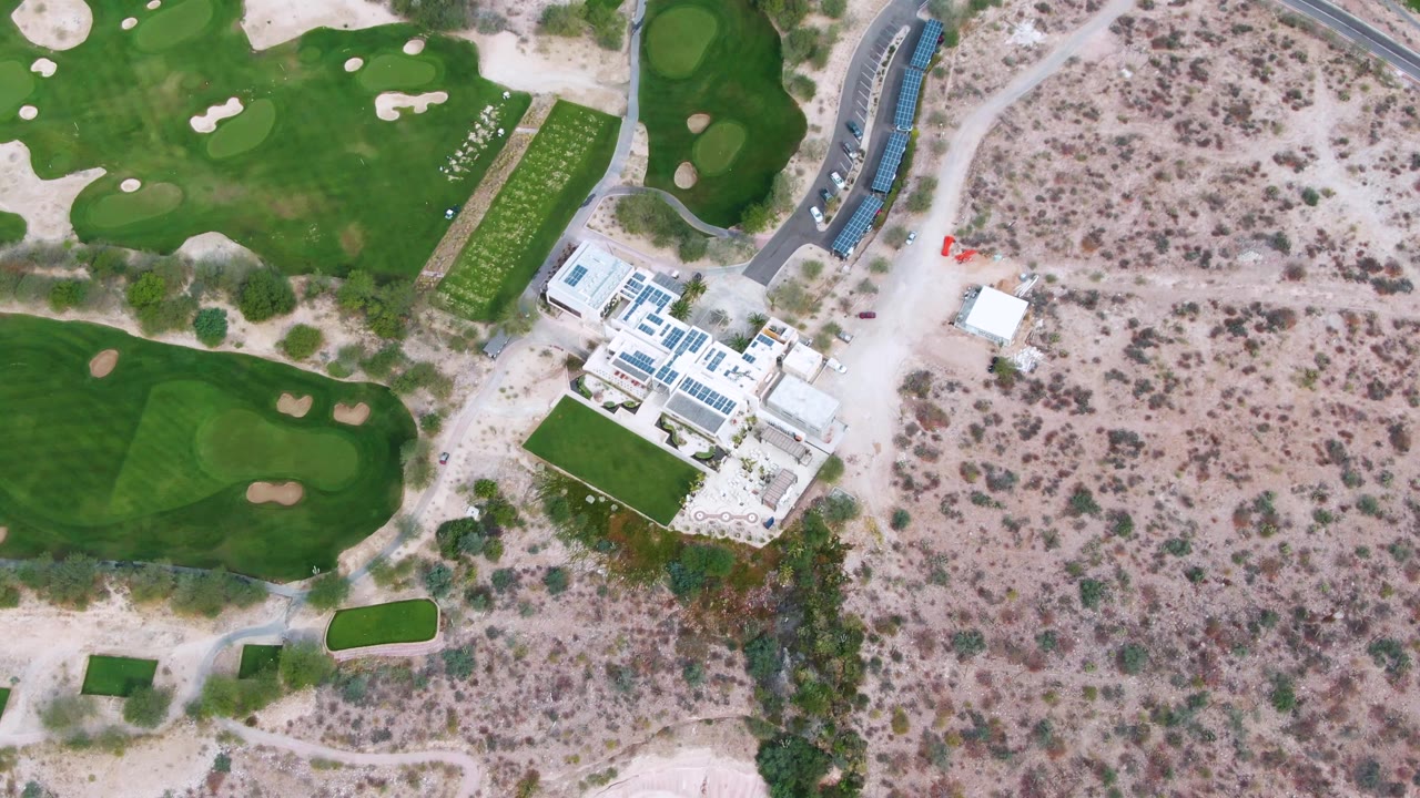 Top-down frame in La Paz, Baja California Sur, showing broad green course areas, bunker shapes, white buildings, a curved road, dry brush, and no visible people.