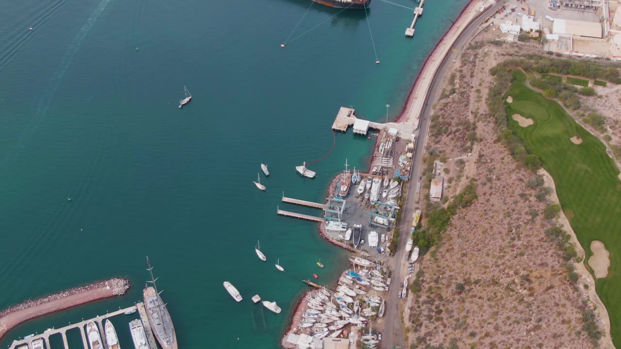 High aerial view of La Paz harbor with small boats spread across turquoise water, docked marina vessels, a curved shoreline road, green grass to the right, a red ship near the top edge, and no visible people.