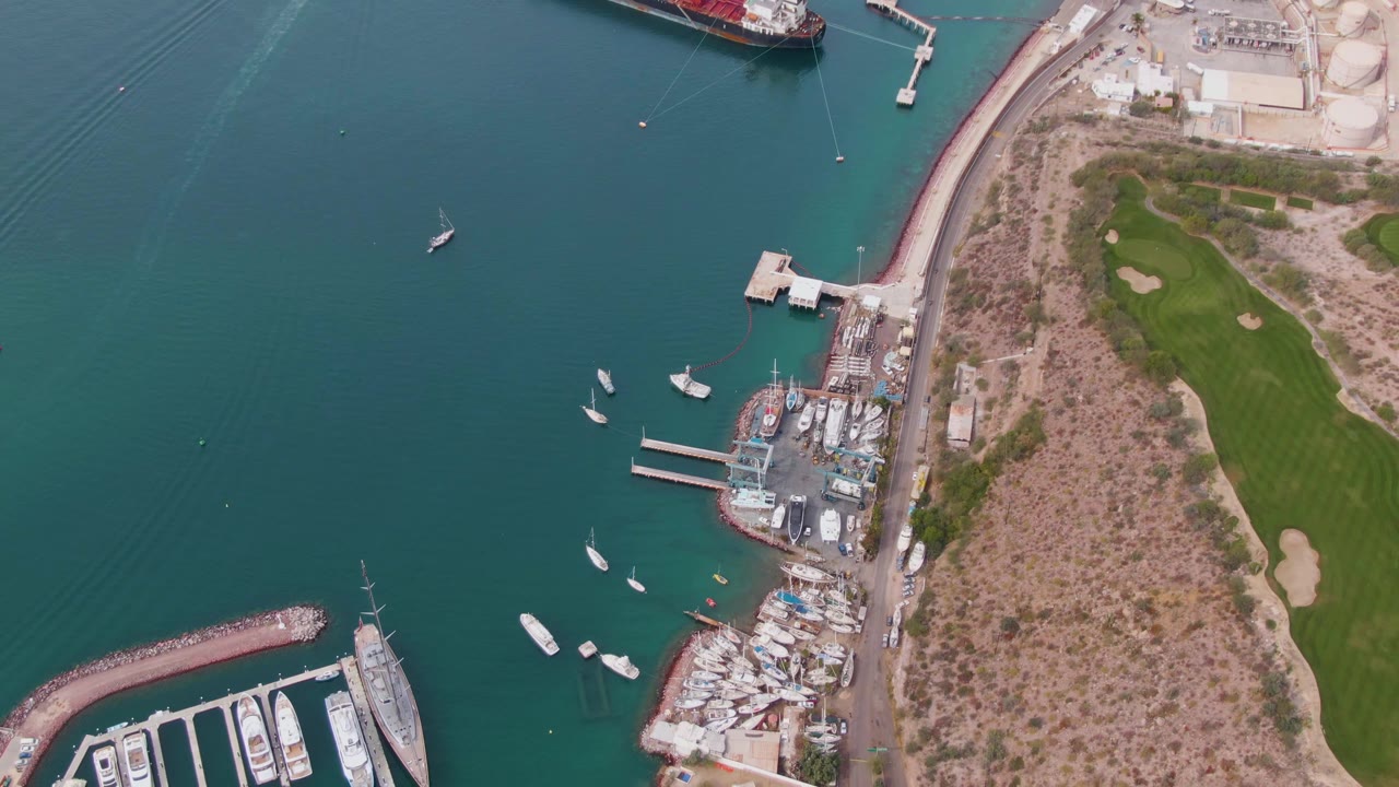 Aerial frame over La Paz marina with a curved breakwater at lower left, multiple boats in the harbor, dock structures along the shore, a road running beside dry terrain, green grass on the right, and no visible people.