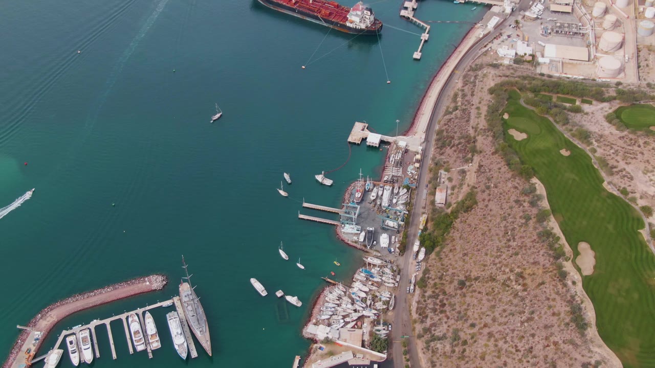 Aerial view of La Paz harbor as a white boat wake enters from the left side of the water, with marina docks and moored boats below, a shoreline road to the right, green grass beyond it, and no visible people.