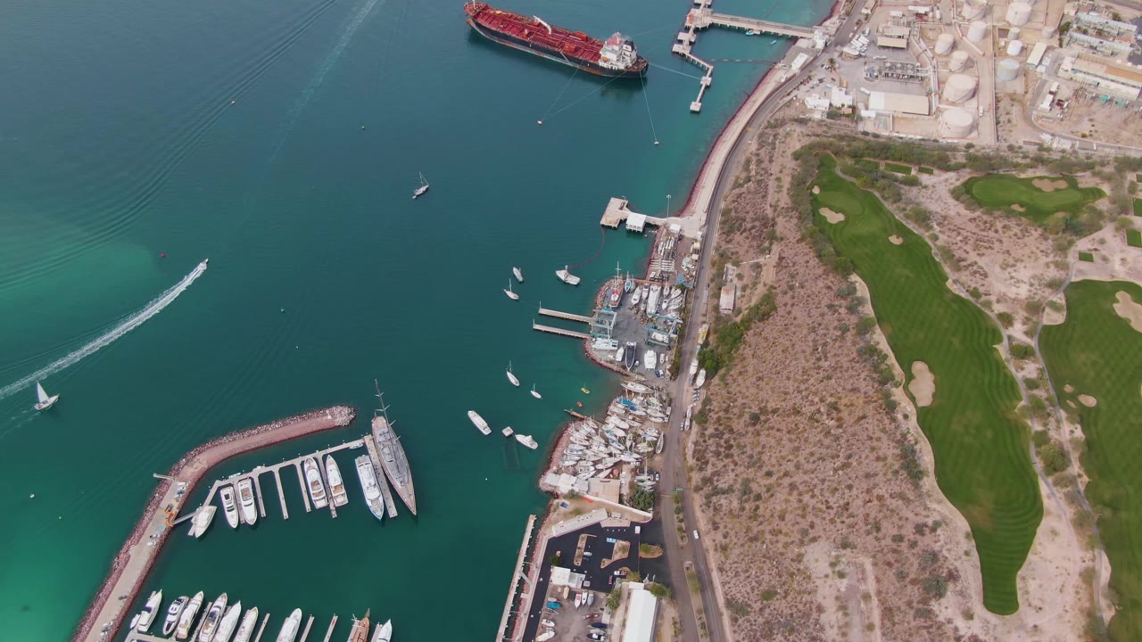 Aerial view over La Paz, Baja California Sur, with teal water, a curved marina breakwater, docked yachts, a shoreline road, green field patches, a red ship near the upper pier, and no visible people.