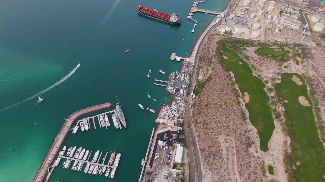 High aerial frame over La Paz showing a diagonal white wake and small sailboat at left, marina slips and yachts below, clustered waterfront structures, coastal road, green fields, and no visible people.