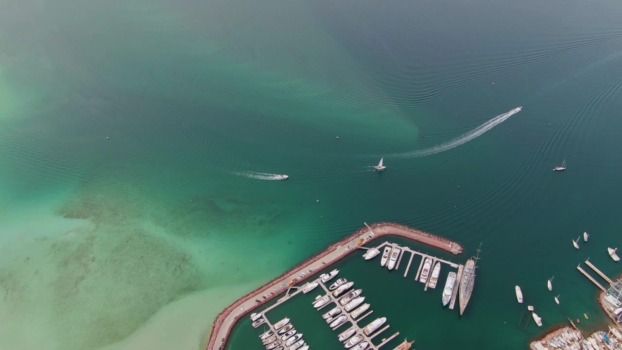 Aerial frame in La Paz, Baja California Sur, with the curved marina breakwater lower in frame, a speedboat leaving a white wake, a sailboat nearby, and no visible people.