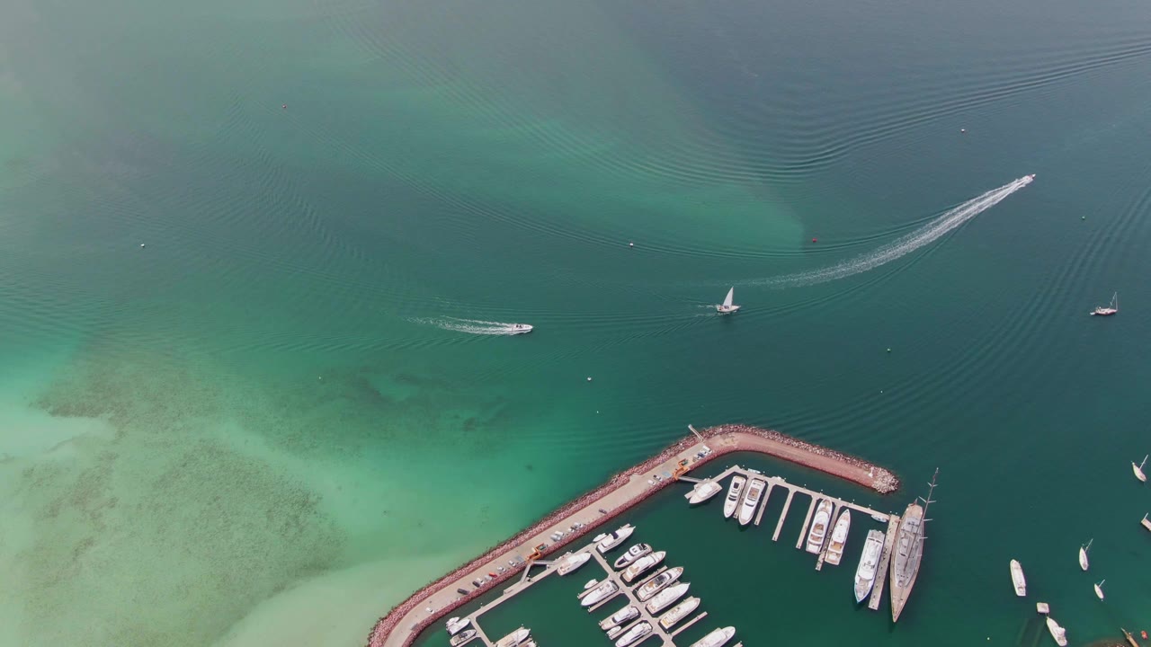 Drone-style overhead frame in La Paz, Baja California Sur, with a sailboat near center, a speedboat wake sweeping across the top, marina docks below, and no visible people.