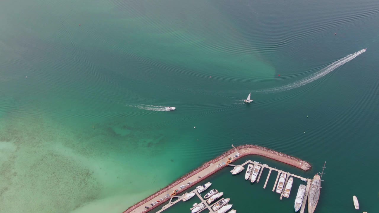 Aerial coastal water with marina corner, sailboat, speedboat, and visible wakes.