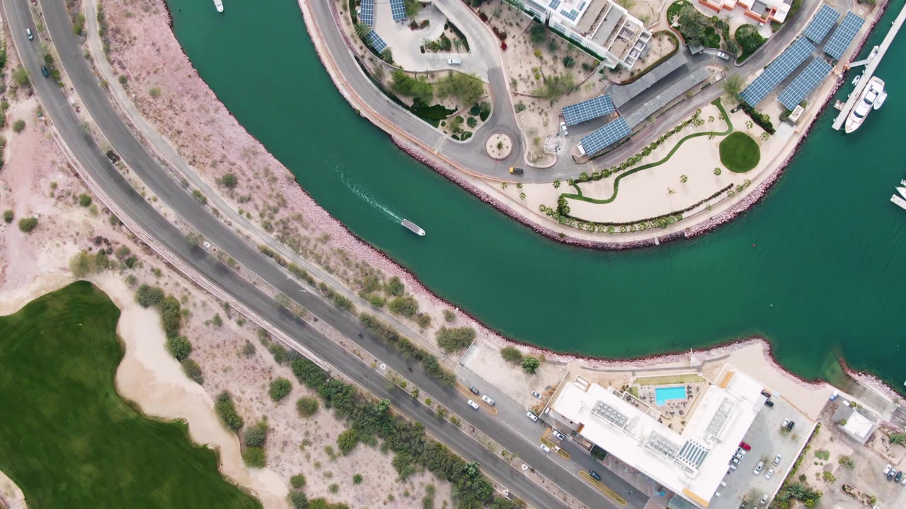 Aerial view in La Paz, Baja California Sur with the small boat farther along the canal, its wake trailing behind, while road vehicles, pale shoreline, buildings, and no visible people remain in view.