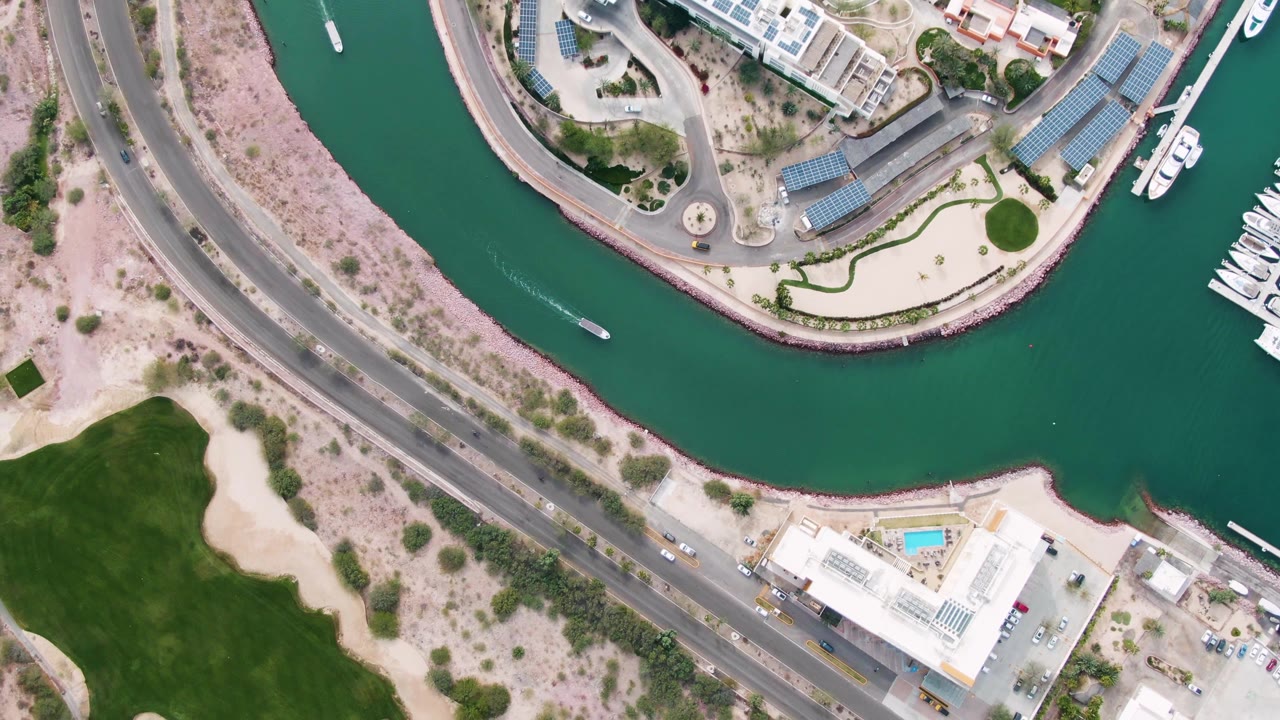 Top-down La Paz marina canal view showing the boat mid-channel, a curved waterfront road, buildings with light roofs, marina docks with white vessels at frame edge, and no visible people.