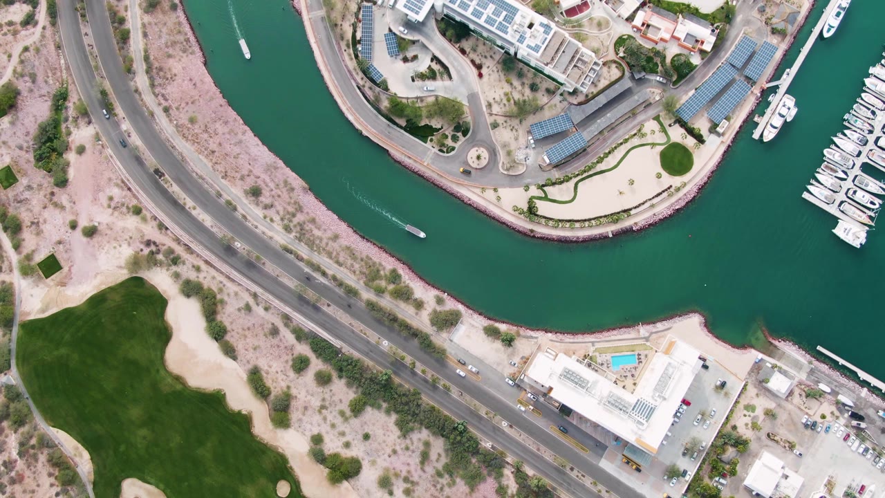 La Paz, Baja California Sur aerial frame with the boat crossing the canal, marina slips and white boats on the right, a road with vehicles on the left, green grass below, and no visible people.