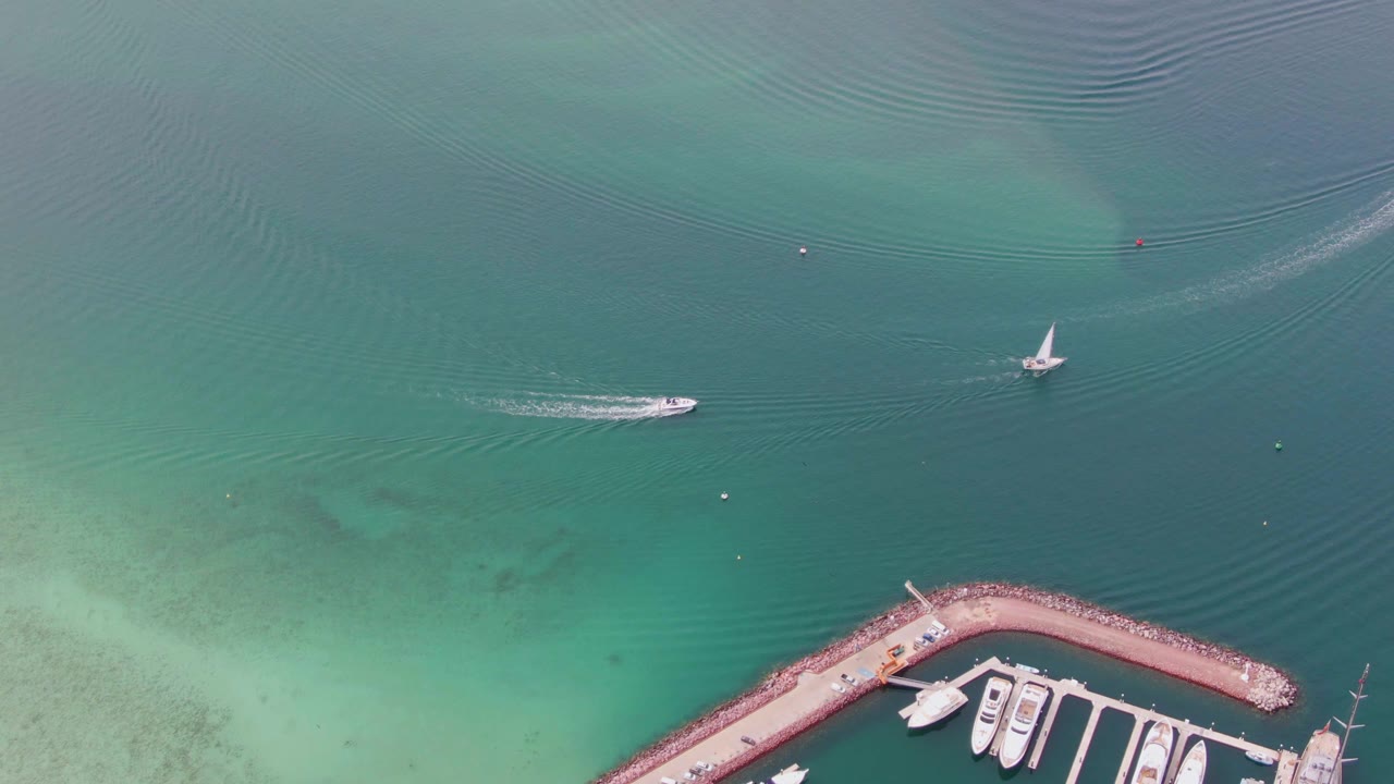 Aerial view in La Paz, Baja California Sur, Mexico with the motorboat and wake shifted along the open water, the sailboat still upright at right, docked boats below, and no visible people.