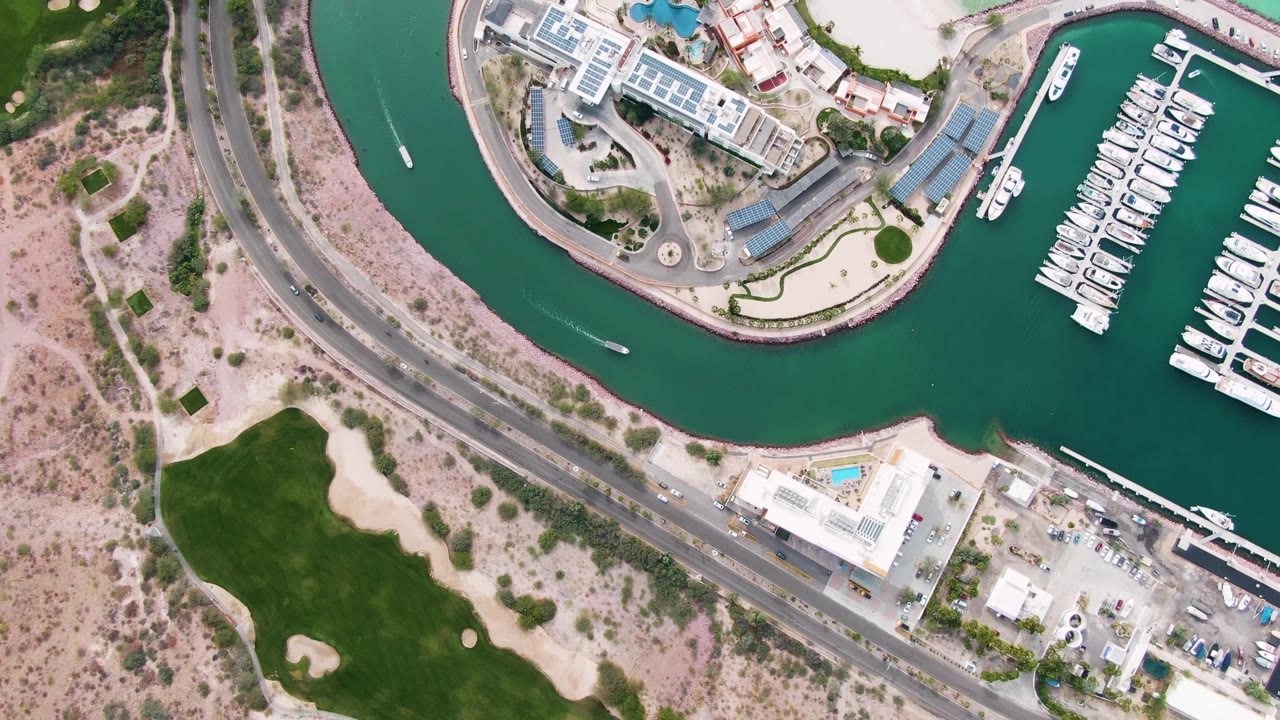 Opening overhead frame of La Paz, Baja California Sur, with teal marina water, docked boats at right, a curved road through the center, golf greens at lower left, and no visible people.