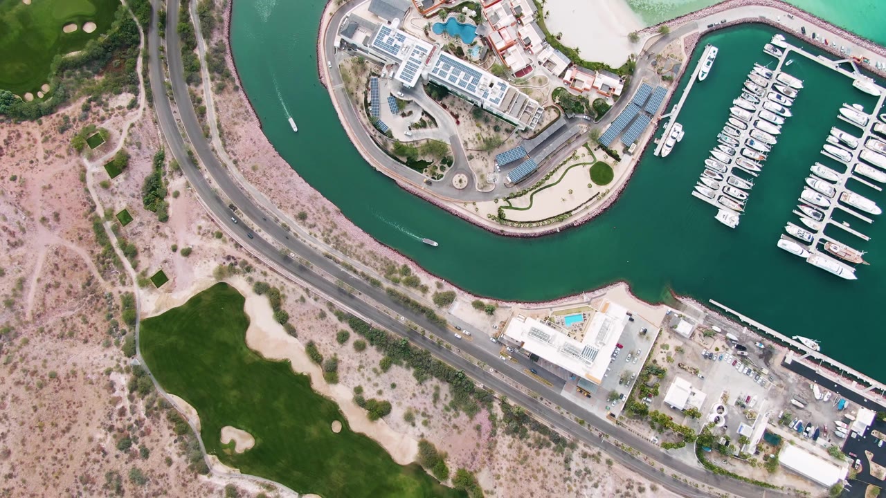 One second into the La Paz aerial, the marina slips and white boats sit farther right while the canal curve, waterfront buildings, central road, and no visible people remain clear.