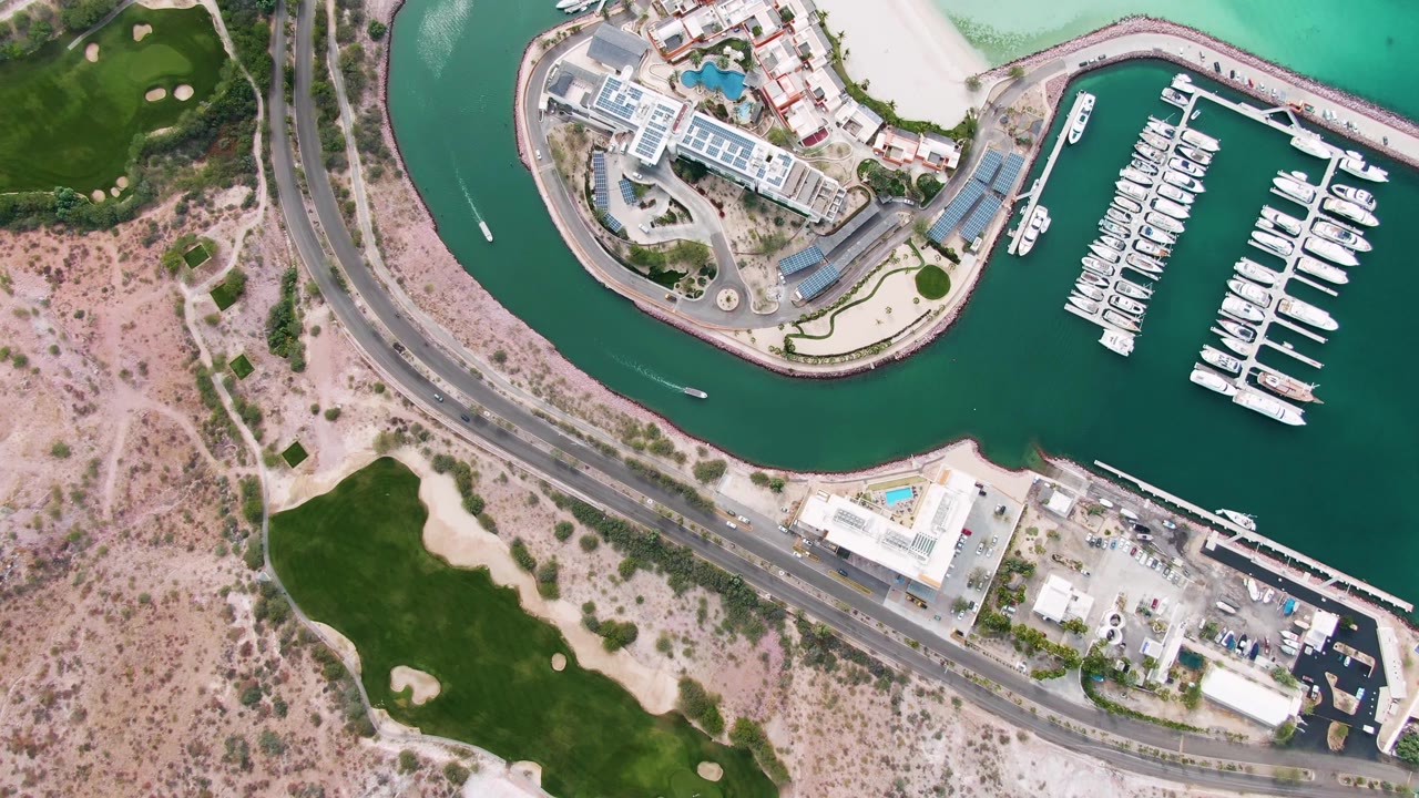 Midway through the clip, the La Paz overhead composition shows docked boats lined in the marina, a sweeping road, waterfront structures, golf fairway edges, desert lots, and no visible people.