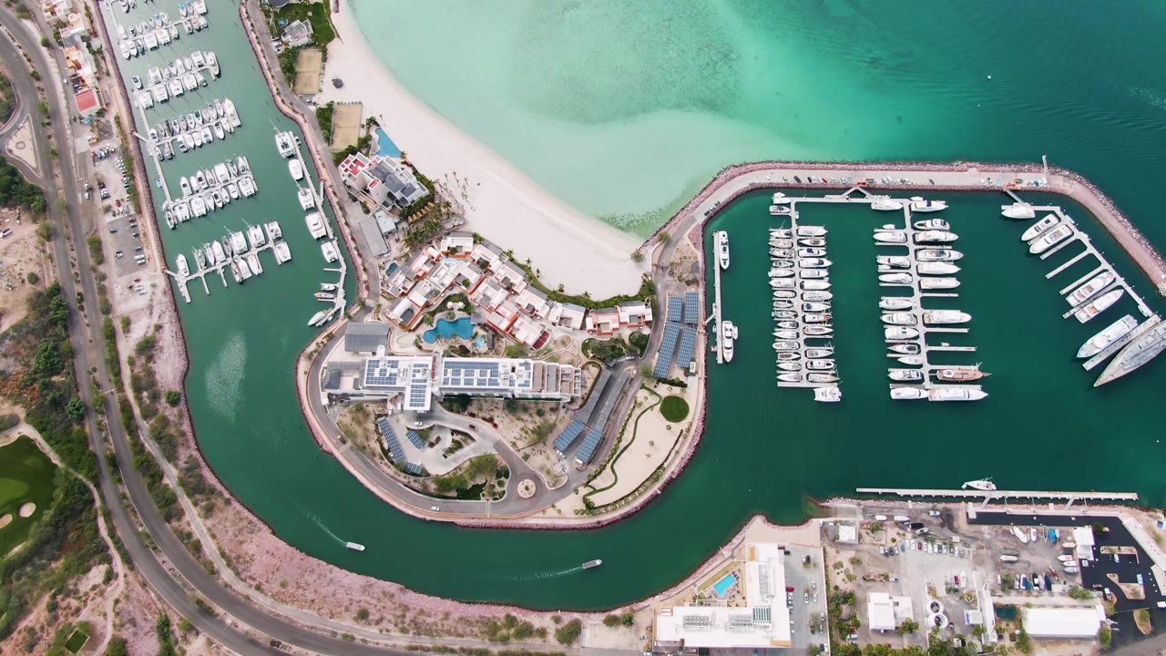 Overhead frame of La Paz, Baja California Sur showing a marina with docked boats, a pale beach, curved road, green-turquoise water, nearby buildings, and no visible people.