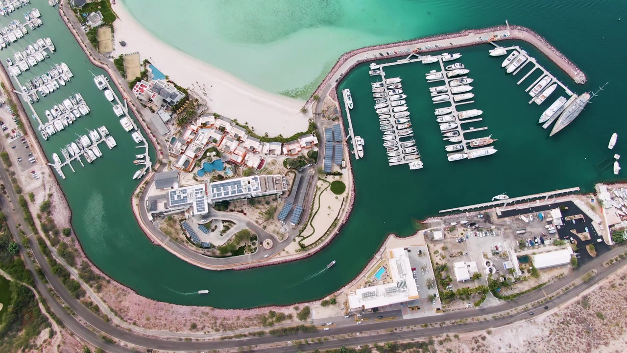 High aerial frame over a La Paz marina with rows of docked boats, pale beach at the top, waterfront buildings and pools, a curved road at the bottom, and no visible people.
