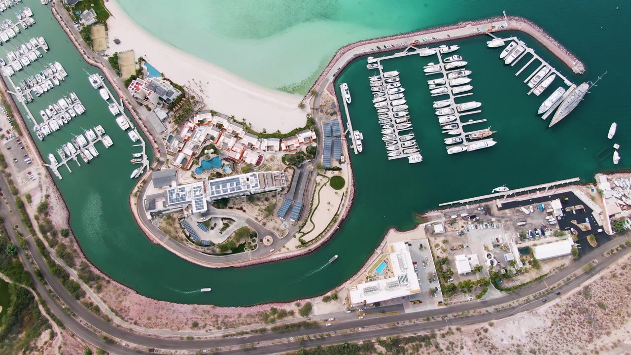 Aerial view of curved marina waterway with boats, beach, buildings, and road.