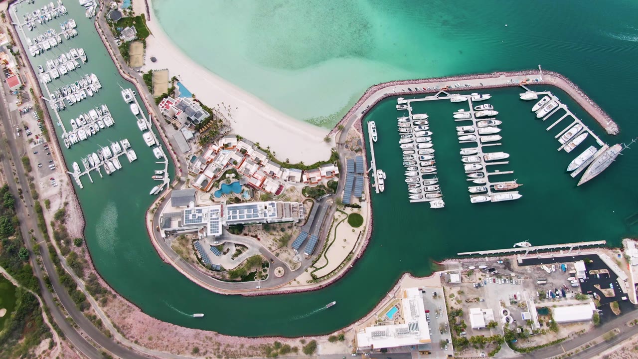 Overhead aerial frame of a La Paz, Baja California Sur marina with white boats at docks, pale sandy beach above, curved green water channel below, surrounding roads and buildings, and no visible people.